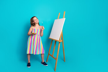 Smiling young girl artist holding paintbrush and palette standing beside easel with blank canvas...
