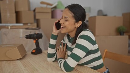Young hispanic woman smiling while talking on phone surrounded by cardboard boxes and a drill inside her new home suggesting a recent relocation or renovation. - Powered by Adobe