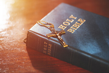 A bronze crucifix necklace resting on a closed Holy Bible, illuminated by warm light, symbolizing...