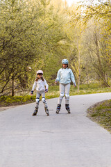 Two girls rollerblading on park path in spring, wearing helmets and knee pads. Children enjoy...