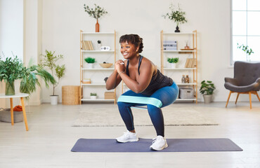Sporty fit middle aged happy african american woman doing squat with elastic band morning exercise...