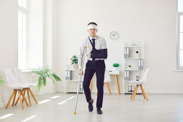 Portrait of a young injured man standing in office with his arm in a plaster cast and bandaged head...