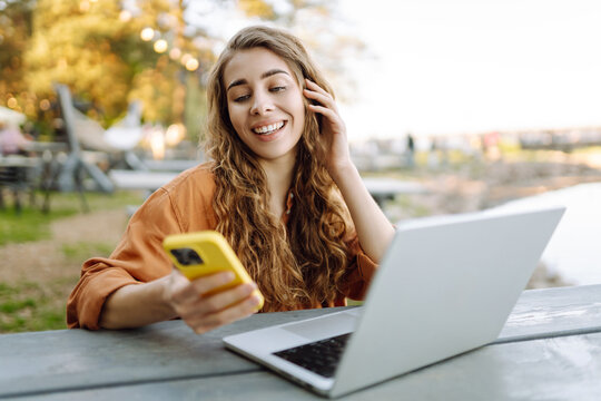Smiling woman with phone and laptop sitting at table in sunny park. Beautiful female freelancer working at laptop at table and chatting on phone. Freelancing concept.