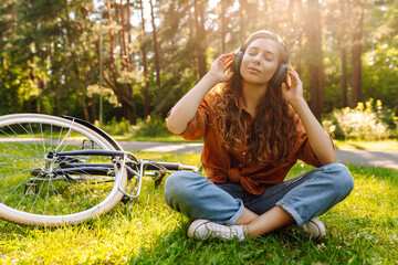 Cheerful woman in wireless headphones relaxing on a green meadow with a bicycle in the background....