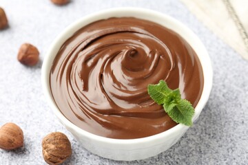 Tasty chocolate spread with mint in bowl and hazelnuts on grey textured table, closeup