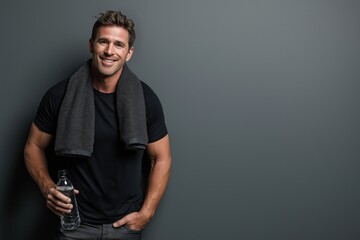 Fototapeta premium A fit man in a black t-shirt with a towel over his shoulders smiles while holding a water bottle against a plain gray background.