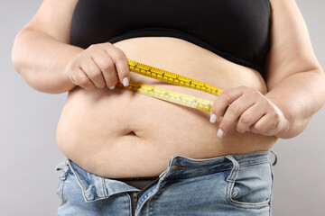 Overweight woman measuring her belly with tape on grey background, closeup