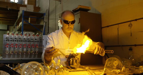 Shaping male glassblower directing flame onto glass tube at studio, with torch and lab coat