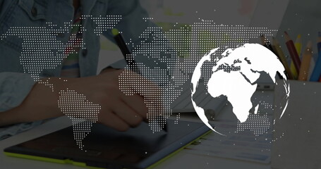 Holding stylus, artist drawing on tablet at desk, showing colored pencils and world map overlay