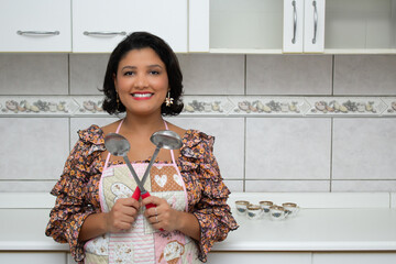 Happy woman in kitchen, holding stainless steel cutlery. Housewife wearing apron and holding large spoons for serving food. Ideal for cooking meals, restaurant chef and gastronomy.