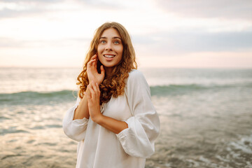 Happy young woman in white dress enjoying sunset rays on seashore. Beautiful woman walking along...