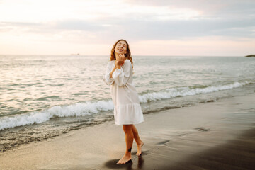 Happy young woman in white dress enjoying sunset rays on seashore. Beautiful woman walking along...