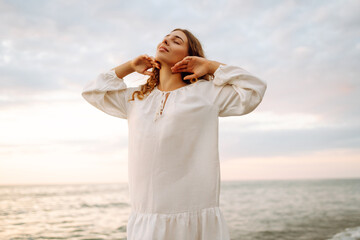 Happy young woman in white dress enjoying sunset rays on seashore. Beautiful woman walking along...