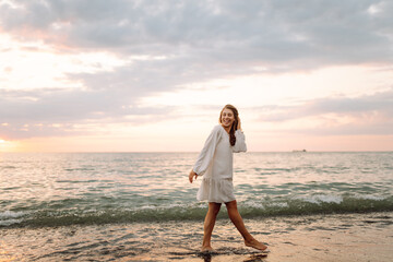 Happy young woman in white dress enjoying sunset rays on seashore. Beautiful woman walking along...