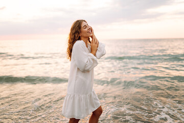 Happy young woman in white dress enjoying sunset rays on seashore. Beautiful woman walking along...
