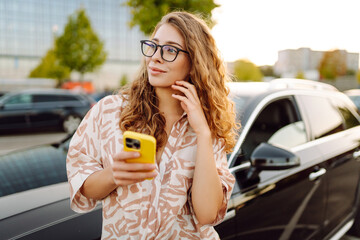 Beautiful woman with curly hair with a phone in her hands stands near a car in a parking lot. Young female driver enjoys the weather and chats at sunset. Transport and technology concept.