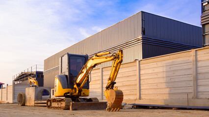Mini excavator on the ground near precast concrete fence wall outside of industrial warehouse factory building structure in construction site, perspective side view