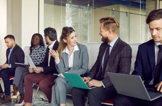 Talking smiling diverse office friendly candidates sitting on chairs in row, business people applicants interns waiting for job interview, holding gadgets, employments and recruitment service at work