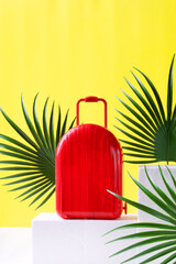 Red suitcase and palm leaves on white podium against the yellow background. Travel and summer vacations concept. Mockup with copy space on the baggage