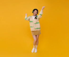Asian woman joyfully dances with headphones on arms raised in excitement wearing a striped cardigan...