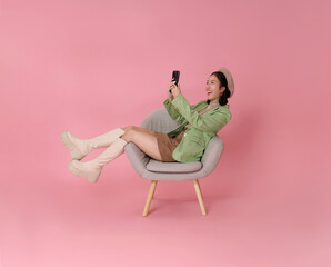 happy Asian woman wearing a green blazer, beret, and boots relaxing in a chair and looking at her phone isolated on pink background.