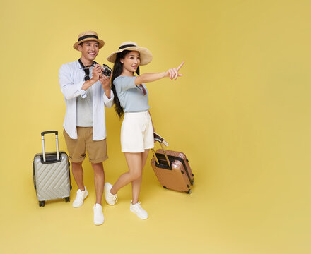 Happy Asian couple with hats luggage and camera pointing into distance Ready for adventure isolated on yellow background. Great for travel, holidays, or exploration themes.