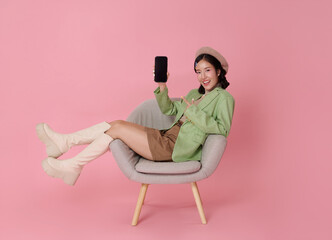 Happy Asian woman sitting in chair showing smartphone with blank white screen isolated on pink background. Ideal for app mockups or ad content.