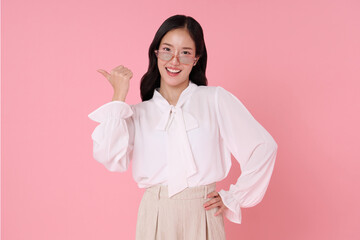 Smiling Asian businesswoman wearing glasses and a white blouse, confidently pointing her thumb behind her with a cheerful expression. Posed against a soft pink background,