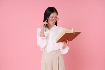 Young Asian woman surprised reading book with glasses and pen isolated on pink background. Expresses discovery amazement.