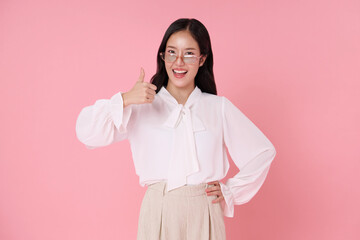 Confident young Asian woman in a white blouse giving a thumbs-up gesture with a cheerful smile, standing against isolated on pink background.