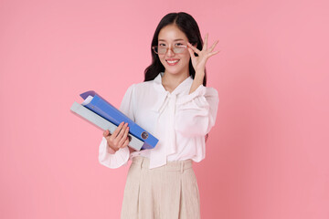 Smiling young Asian businesswoman wearing a white blouse and glasses, holding colorful document binders while posing confidently against isolated on pink background.