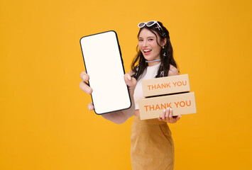 Happy young Asian woman smiling holding package parcel box and showing mobile phone application post mockup isolated on yellow background.