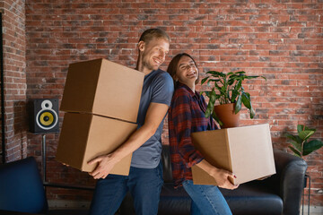 A joyful couple carries boxes and a plant, settling into their new residence