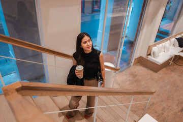 Young manager descending the stairs in a modern office building, carrying a coffee cup and a jacket