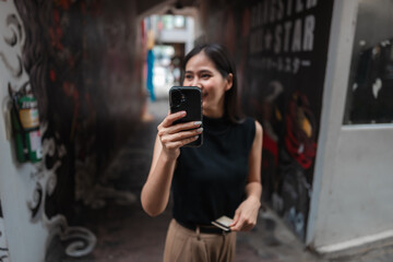 Smiling young woman holding smartphone and credit card making online payment in urban alley with graffiti