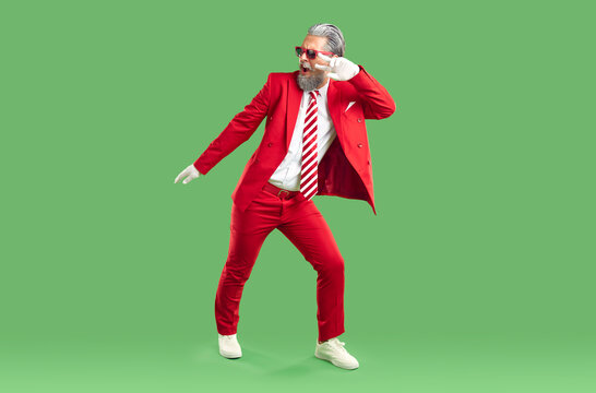 Positive extravagant gray-haired Santa Claus dressed in classic stylish red suit. Studio photo of funny happy Santa Claus fooling around and dancing with his hand raised on isolated green background.