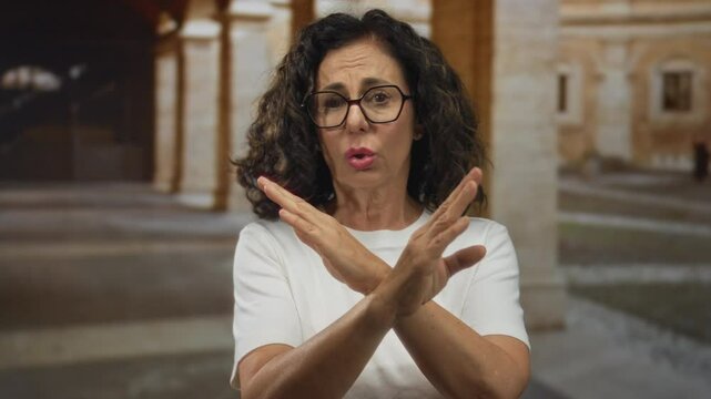 Middle aged hispanic woman crosses bare arms in x gesture of denial in old town stone courtyard; rejection refusal.