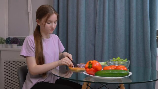 Teenager girl is carefully chopping red onions on a wooden cutting board, preparing a healthy salad with fresh lettuce, cucumber, and bell peppers on a glass table in a home kitchen