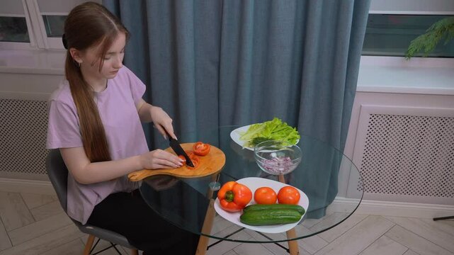 Teen girl neatly chopping tomatoes on a wooden cutting board, surrounded by other fresh vegetables such as lettuce, cucumbers, bell peppers and onions, preparing a healthy salad in the home kitchen