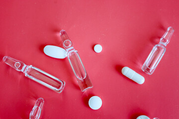 Pharmaceutical ampoules and white pills are scattered across a bright red surface. This composition...