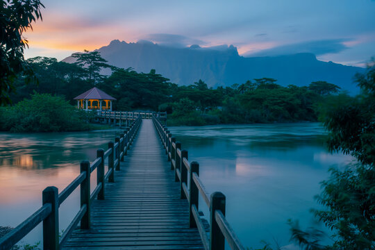 Twilight Serenity Wooden Boardwalk Over Mirror-Like River with Lit Gazebo, Reflecting Pastel Skies and Mountain Silhouettes