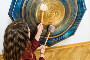 Woman using mallets to play a large gong, creating soothing sounds for a sound bath session. The...