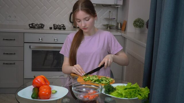 Young teen girl neatly slicing cucumber on glass table, preparing fresh, nutritious salad with colorful vegetables in bright home kitchen workplace