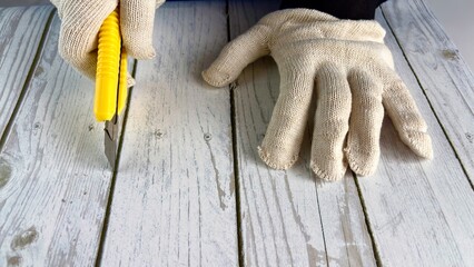 Gloved hands cut a wooden surface with a bright yellow utility knife