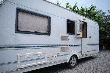 A white camper trailer is parked on a paved backyard near a brick house and cherry tree. RV travel content, camping promotions, suburban lifestyle visuals, or articles about alternative housing