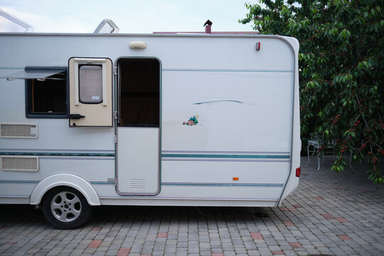 A white camper trailer is parked on a paved backyard near a brick house and cherry tree. RV travel content, camping promotions, suburban lifestyle visuals, or articles about alternative housing