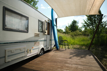 A camper van with a wooden terrace and fabric canopy is parked in the middle of tall grass and trees. This peaceful scene evokes a sense of freedom, glamping, and off-grid living.
