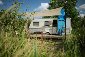 A camper van with a wooden terrace and fabric canopy is parked in the middle of tall grass and trees. This peaceful scene evokes a sense of freedom, glamping, and off-grid living.