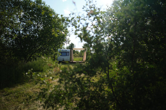 A camper van with a wooden terrace and fabric canopy is parked in the middle of tall grass and trees. This peaceful scene evokes a sense of freedom, glamping, and off-grid living.