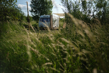 A camper van with a wooden terrace and fabric canopy is parked in the middle of tall grass and trees. This peaceful scene evokes a sense of freedom, glamping, and off-grid living.